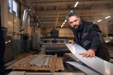 Portrait of man in protective clothes and glasses standing near woodworking machine and cutting material in workshop