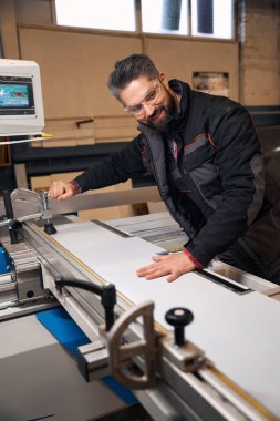 Portrait of smiling man in protective clothes and glasses standing near woodworking machine and cutting material in workshop