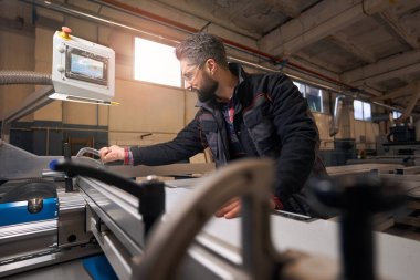 Portrait of smiling male in protective clothes and glasses standing near woodworking machine and cutting material