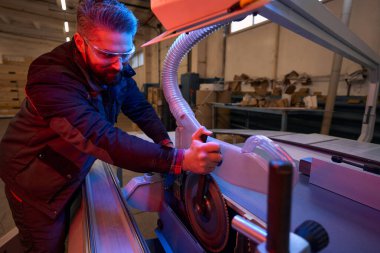 Worker in protective clothes and glasses standing near form cutting machine and repairing it