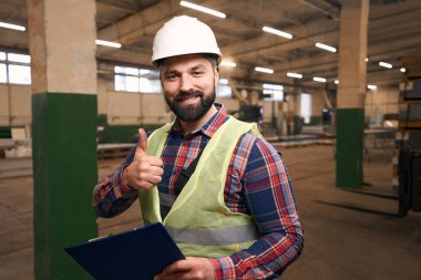 Smiling man in protective clothes and helmet, standing in workshop, holding clipboard and showing gesture agree