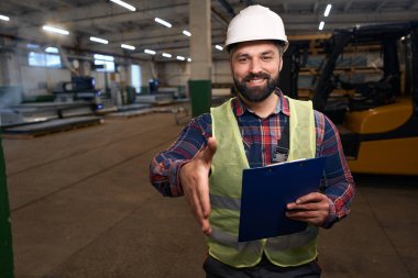 Smiling male in protective clothes and helmet, standing in workshop, holding clipboard and extend hand to shake