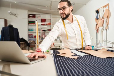 Calm concentrated tailor standing at cutting table and looking at laptop screen