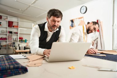 Smiling tailor pinning pattern pieces on fabric while his focused coworker typing on laptop