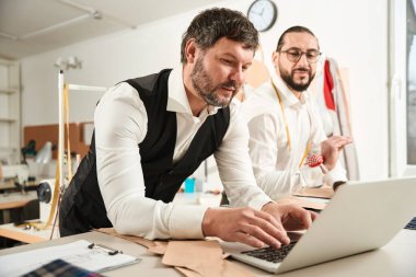 Concentrated fashion designer working on portable computer in presence of his colleague