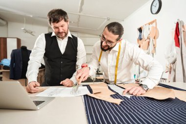 Smiling couturier leaning on cutting table and pointing with pair of scissors at suit sketch to colleague