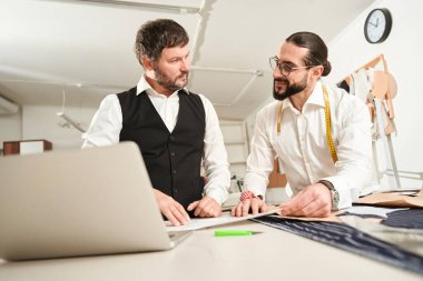 Smiling young tailor and his colleague standing at cutting table with paper patterns laid out on fabric