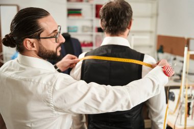 Calm concentrated young Caucasian tailor measuring client shoulder width in his studio