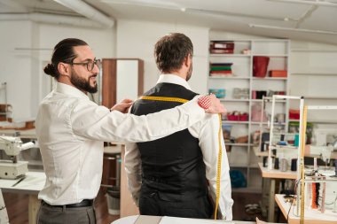 Focused couturier measuring customer shoulder width using tape measure in his atelier