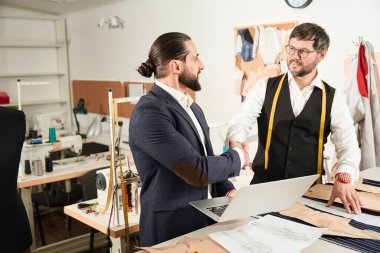 Smiling pleased fashion designer shaking hands with his colleague at cutting table