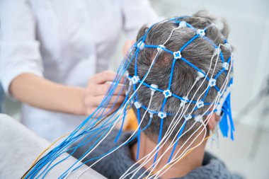 Patient in a hospital gown sits in a chair with sensors on his head, a medical worker controls the process