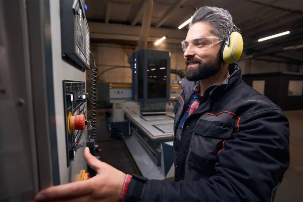 Worker in protective clothes, glasses and anti-noise headphones standing in workshop near equipment, selecting command on control panel