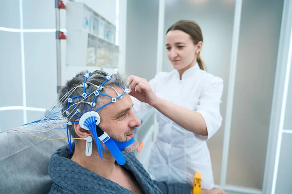 Man in a hospital gown undergoes a check-up in hospital, a young diagnostician performs EEG procedure for him - electroencephalography