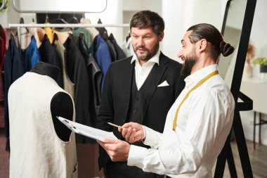 Joyous tailor pointing with pencil at sheet of paper with sketch to his client