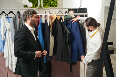 Serious concentrated gentleman looking at himself in mirror in presence of busy tailor