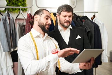 Waist-up portrait of pleased tailor demonstrating something to client on tablet computer