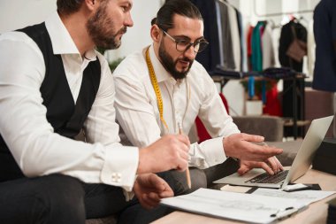 Fashion designer seated at table pointing at laptop screen to his colleague