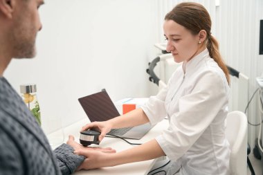 Waist up side view portrait of focused elegant Caucasian doctor is using oligoscan for examining body indicators in medicine clinic