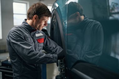 Man in protective clothes standing in tire fitting near automobile and checking front wheel bearing