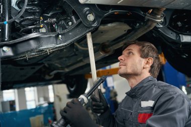 Man in protective clothes standing in tire fitting under car and inspecting rear wheel independent suspension arm