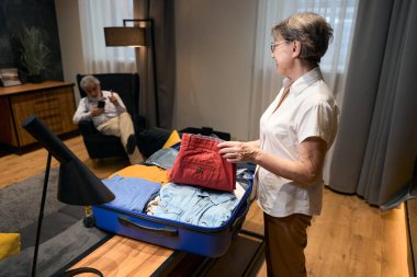 Elderly female standing in hotel, putting clothes in suitcase, male sitting and looking at phone