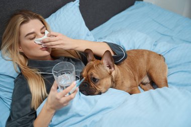 Ill woman holding digital thermometer and glass of water while blowing nose into handkerchief