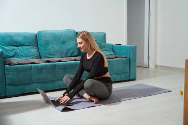 Smiling pleased athletic female sitting cross-legged on yoga mat typing on portable computer