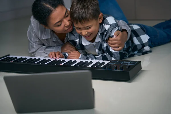African american mother hugging her teenage son shoulders, they sit comfortably on the floor with a digital piano and laptop