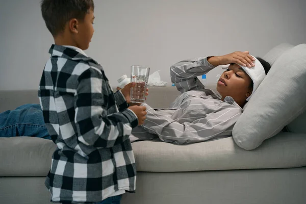 Boy brought a glass of water to his sick mother, the woman lies on the sofa with a headache