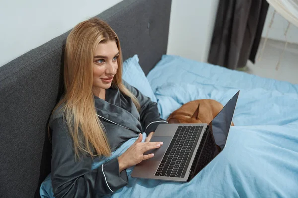 Smiling dreamy young woman seated in bed with portable computer and dog
