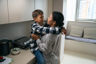 Nice african american female is resting at home in the kitchen with her son, the boy gently hugs his mother