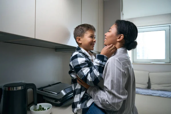 Smiling african american mom and son settled down at home in the kitchen, the boy gently hugs mom