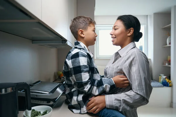 Funny african american mom and son settled down at home in the kitchen, the boy tickles mom