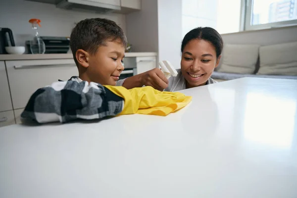 Child in a plaid shirt and protective gloves washes the kitchen surface, next to it is a happy mother with a spray bottle