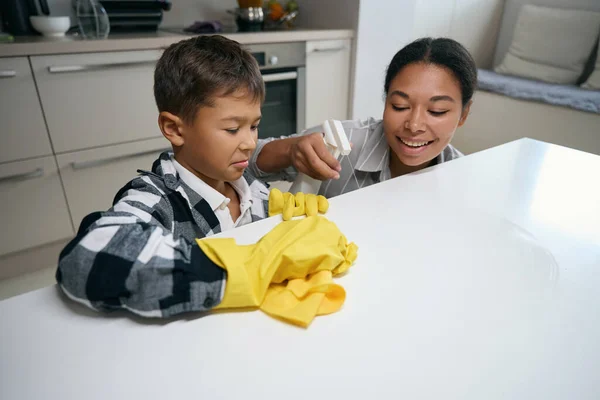Boy in a plaid shirt and protective gloves washes the kitchen surface, a smiling mother uses a spray bottle
