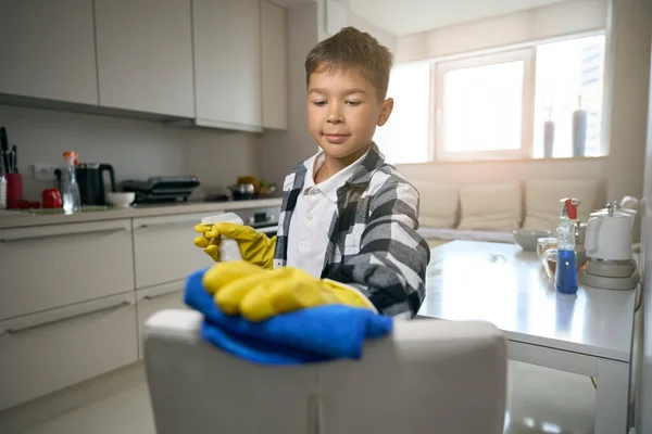 Boy in protective gloves helps in cleaning the kitchen, he uses a rag and a spray bottle