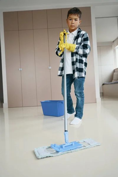 Neat boy washes the floor in the living room with a mop, a teenager in clean casual clothes