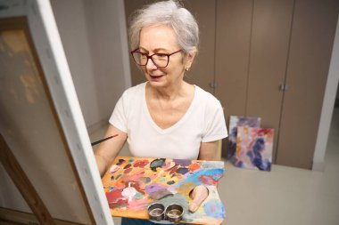 Pensioner holds a palette for drawing in her hands, she stands in front of an easel