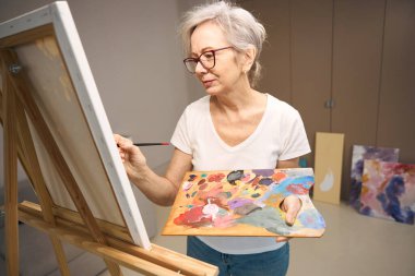 Elderly woman in casual clothes stands in front of an easel, she paints a picture