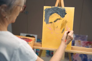 Gray-haired woman paints a portrait with paints and a brush, drawing her hobby
