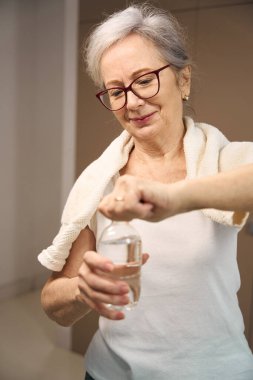 Gray-haired pleasant woman opens a bottle of water, the woman has a towel on her shoulders
