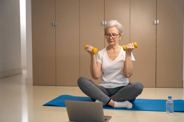 Elderly woman sits with dumbbells in her hands on a karimate, in front of her is a laptop clipart