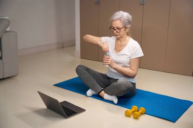 Older female sits on a karimat in a lotus position, in front of her is a laptop and dumbbells