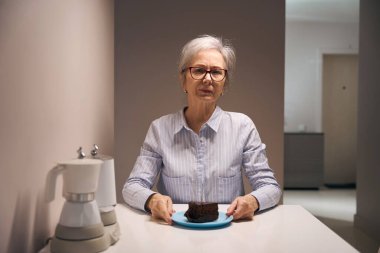 Charming elderly lady is sitting at the table with piece of cake, on the table is set of electric kettles