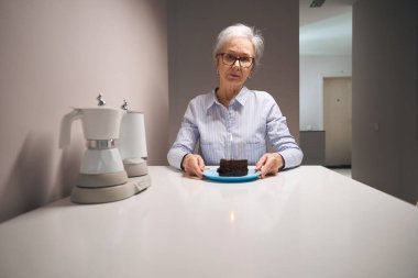 Lonely pensioner sits at a table with piece of cake, on the table there is a set of electric kettles
