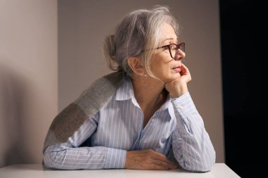 Intelligent elderly woman sits at a table at home with a shawl on her shoulders, she has a beautiful profile