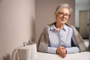 Pretty elderly woman sits at home at the kitchen table, she has a shawl on her shoulders