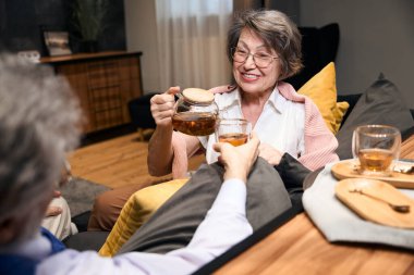Elderly male and woman sitting on sofa in hotel, man holding cup, female pouring drink