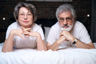 Elderly happy lady and male lying on bed in bedroom, looking at the camera