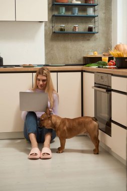 Serious focused woman seated on floor working on laptop in presence of her French bulldog